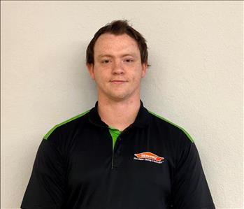 Photo of white male, with clean cut dark hair, dressed in black and green SERVPRO logo polo shirt against cream colored wall